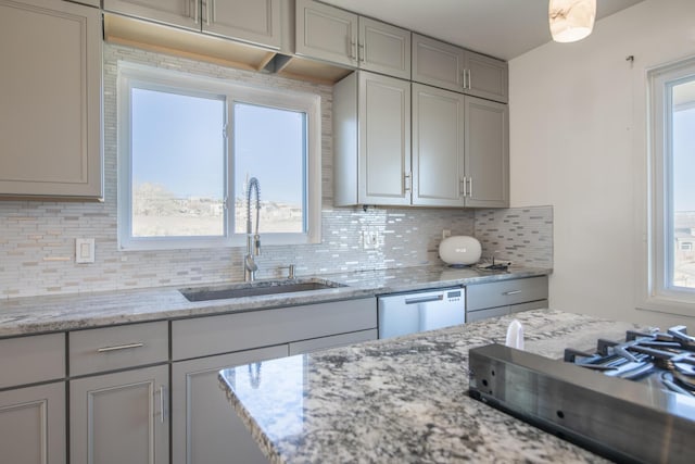 kitchen featuring light stone counters, tasteful backsplash, gray cabinets, stainless steel dishwasher, and a sink