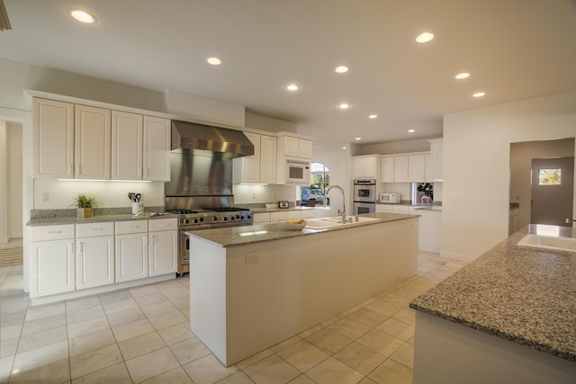 kitchen with wall chimney exhaust hood, white cabinetry, stone countertops, appliances with stainless steel finishes, and a kitchen island with sink