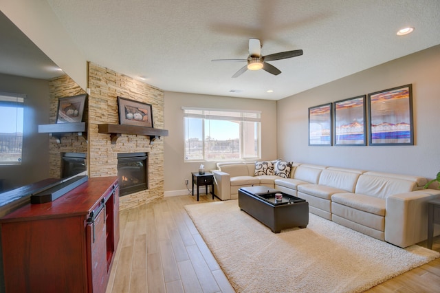 living room featuring ceiling fan, a stone fireplace, light wood-type flooring, and a textured ceiling