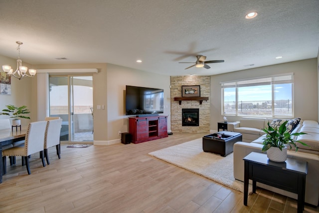 living room featuring ceiling fan with notable chandelier, light hardwood / wood-style floors, a stone fireplace, and a textured ceiling