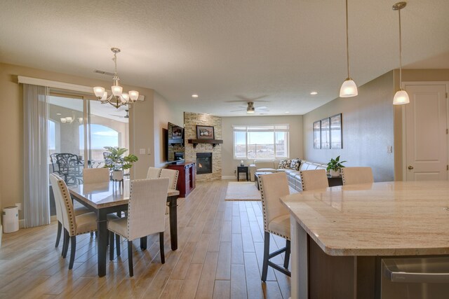 dining room featuring a textured ceiling, ceiling fan with notable chandelier, light hardwood / wood-style flooring, and a stone fireplace