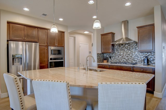 kitchen with a kitchen island with sink, wall chimney range hood, sink, light wood-type flooring, and appliances with stainless steel finishes