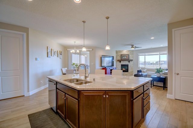 kitchen with light wood-type flooring, stainless steel dishwasher, sink, a stone fireplace, and an island with sink