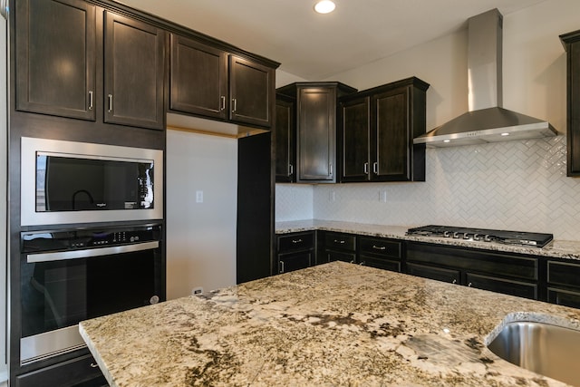 kitchen featuring light stone countertops, tasteful backsplash, dark brown cabinets, stainless steel appliances, and wall chimney range hood