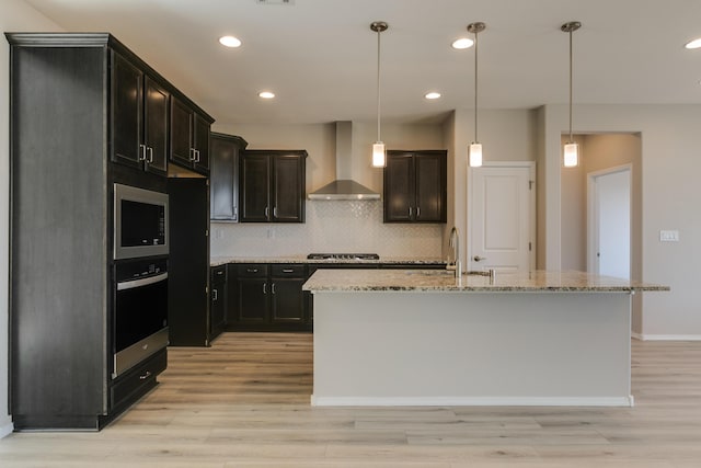 kitchen featuring a center island with sink, sink, wall chimney exhaust hood, light stone counters, and stainless steel appliances