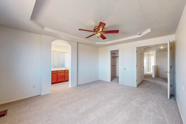 unfurnished bedroom with ceiling fan, ensuite bathroom, light colored carpet, a tray ceiling, and a walk in closet