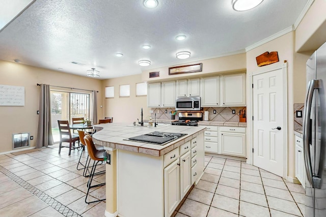 kitchen with decorative backsplash, appliances with stainless steel finishes, light tile patterned floors, tile counters, and an island with sink