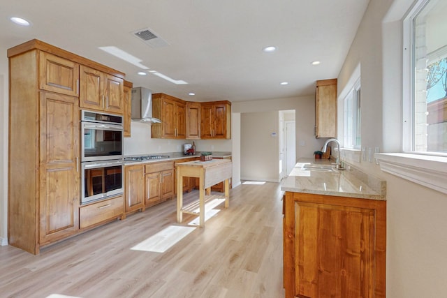 kitchen with wall chimney range hood, sink, light hardwood / wood-style flooring, light stone countertops, and stainless steel appliances