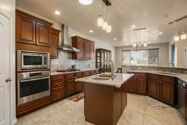 kitchen featuring decorative light fixtures, sink, a kitchen island with sink, stainless steel appliances, and wall chimney exhaust hood