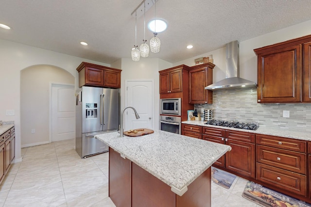 kitchen with appliances with stainless steel finishes, pendant lighting, a kitchen island with sink, decorative backsplash, and wall chimney range hood