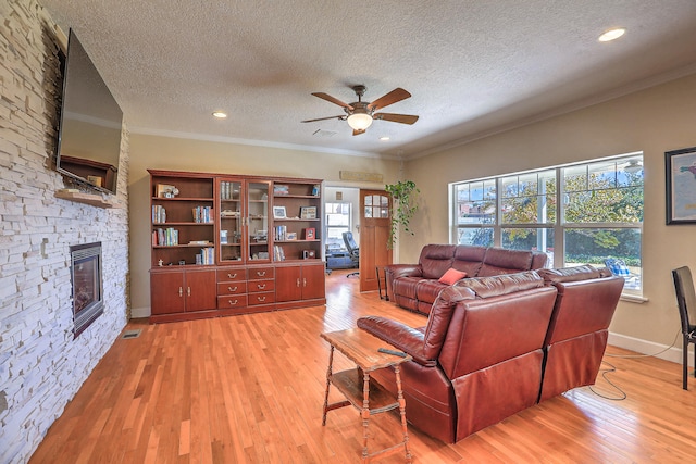 living room with ceiling fan, a stone fireplace, light hardwood / wood-style floors, a textured ceiling, and ornamental molding