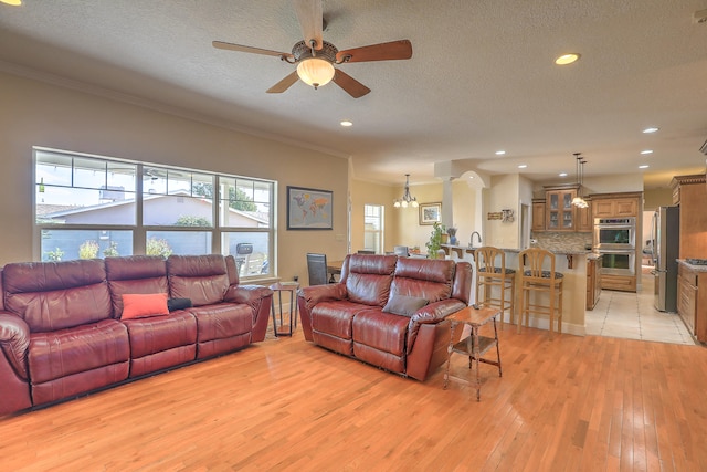 living room with decorative columns, ornamental molding, a textured ceiling, ceiling fan with notable chandelier, and light hardwood / wood-style flooring