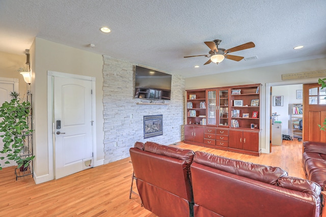 living room with a textured ceiling, light wood-type flooring, a stone fireplace, and ceiling fan