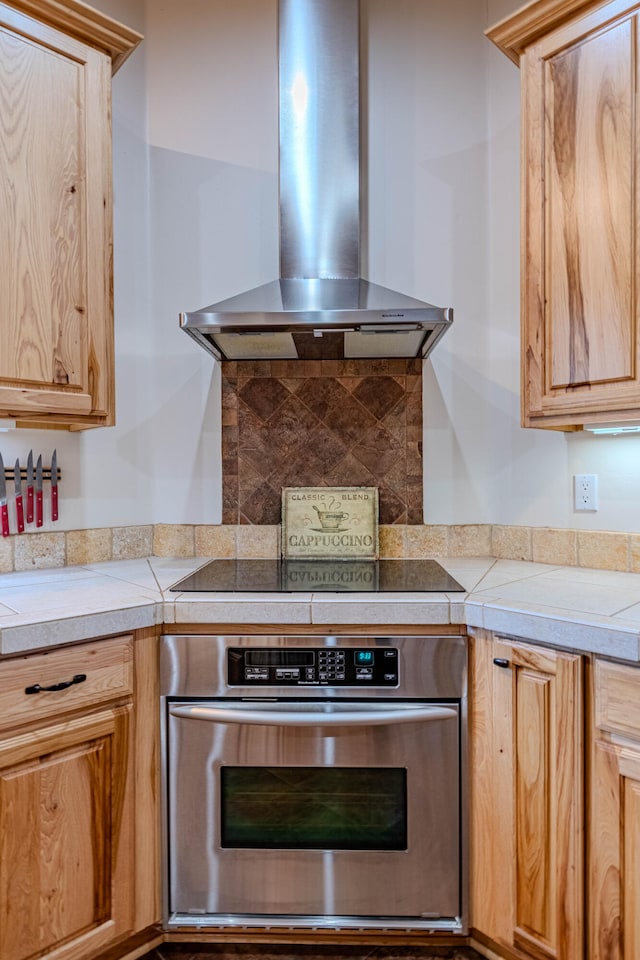 kitchen with decorative backsplash, black electric stovetop, wall chimney exhaust hood, stainless steel oven, and tile countertops