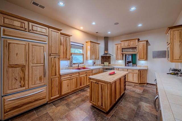 kitchen with wall chimney range hood, stainless steel stovetop, paneled built in fridge, a kitchen island, and tile counters