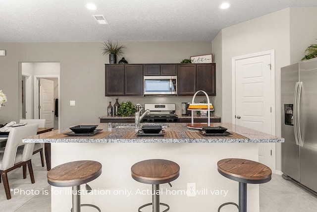 kitchen featuring a breakfast bar, a center island with sink, a textured ceiling, dark brown cabinets, and stainless steel appliances