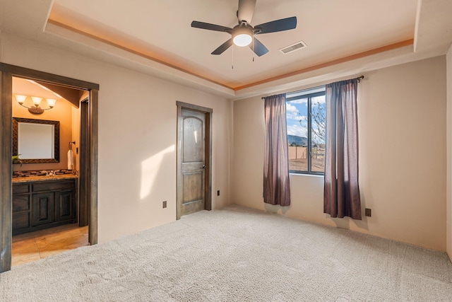 unfurnished bedroom featuring ceiling fan, a raised ceiling, ensuite bathroom, and light carpet