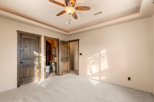 unfurnished bedroom with light carpet, ensuite bathroom, a raised ceiling, and ceiling fan