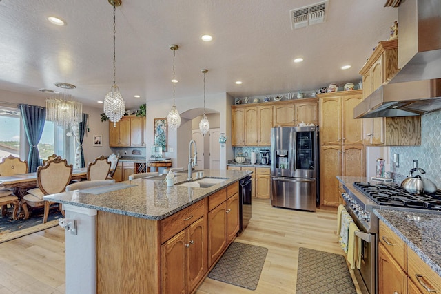 kitchen featuring a sink, visible vents, appliances with stainless steel finishes, wall chimney exhaust hood, and a center island with sink