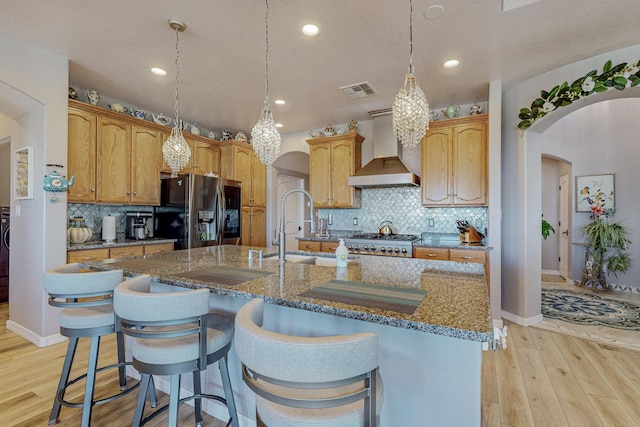 kitchen with a center island with sink, arched walkways, stainless steel fridge with ice dispenser, wall chimney range hood, and a sink