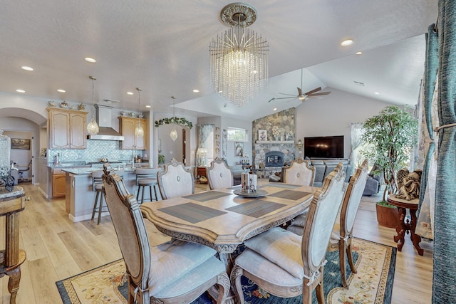 dining space with light wood-style floors, arched walkways, vaulted ceiling, and a stone fireplace