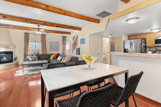 dining area with beamed ceiling, ceiling fan, a large fireplace, and light hardwood / wood-style flooring