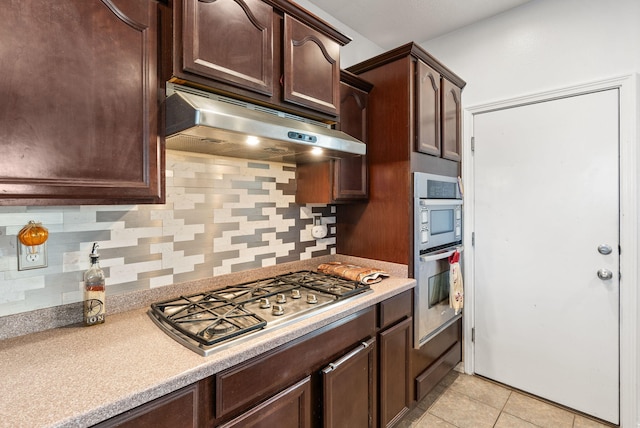 kitchen featuring dark brown cabinetry, stainless steel appliances, backsplash, light tile patterned floors, and exhaust hood