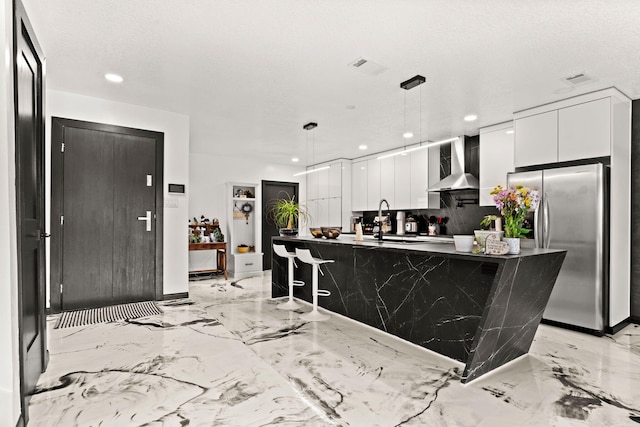 kitchen featuring wall chimney range hood, white cabinetry, an island with sink, and decorative light fixtures