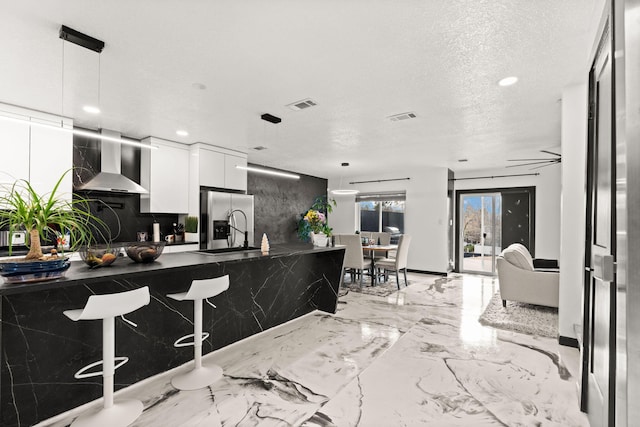 kitchen with hanging light fixtures, wall chimney exhaust hood, a textured ceiling, white cabinetry, and stainless steel fridge with ice dispenser