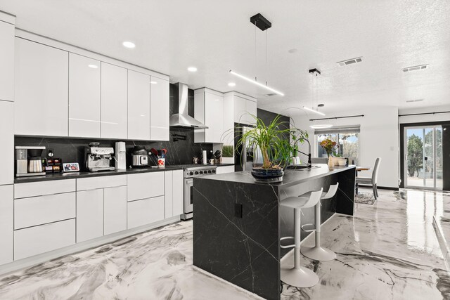 kitchen featuring wall chimney range hood, tasteful backsplash, an island with sink, decorative light fixtures, and white cabinets