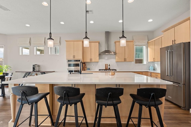 kitchen featuring wall chimney exhaust hood, light brown cabinets, a kitchen island with sink, and appliances with stainless steel finishes