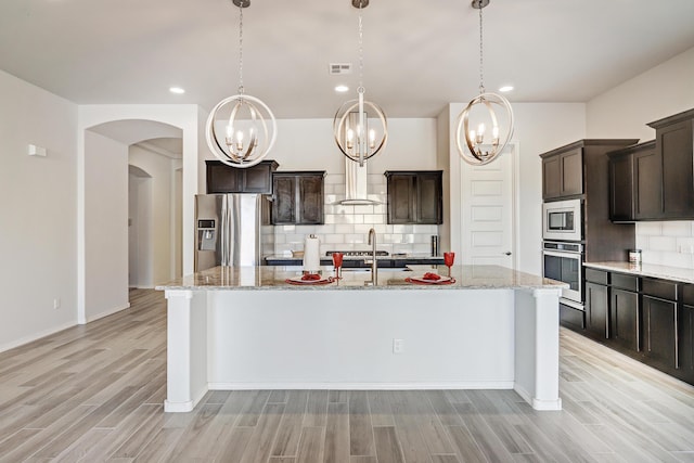 kitchen with appliances with stainless steel finishes, decorative light fixtures, backsplash, dark brown cabinets, and a center island with sink