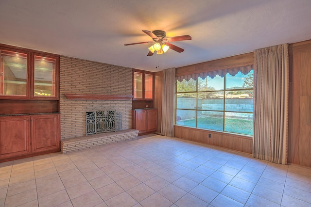 unfurnished living room with ceiling fan, a brick fireplace, a textured ceiling, wooden walls, and light tile patterned floors