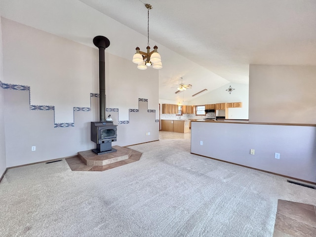 unfurnished living room with ceiling fan with notable chandelier, a wood stove, light carpet, and vaulted ceiling