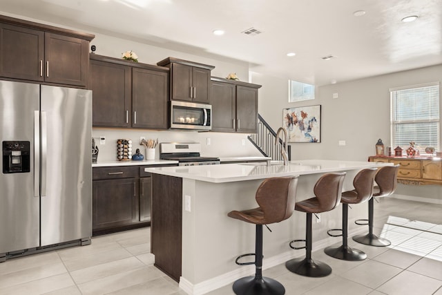 kitchen featuring dark brown cabinetry, a breakfast bar, light tile patterned floors, appliances with stainless steel finishes, and an island with sink