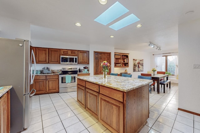 kitchen with rail lighting, stainless steel appliances, a skylight, light stone counters, and a kitchen island