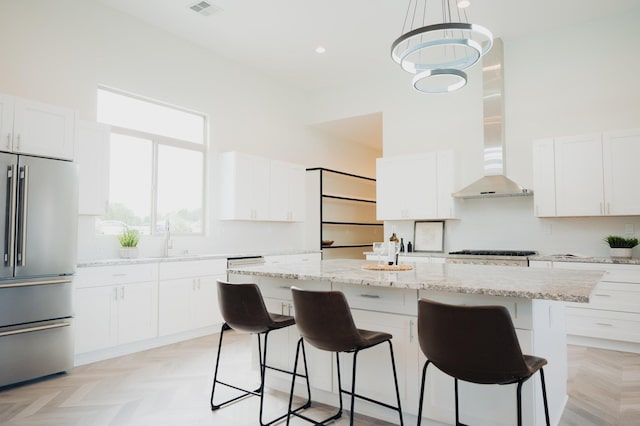 kitchen with a center island, wall chimney range hood, decorative light fixtures, white cabinets, and appliances with stainless steel finishes