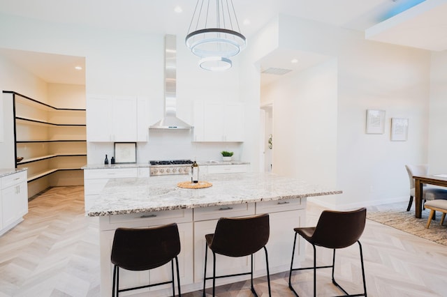 kitchen featuring white cabinets, hanging light fixtures, wall chimney exhaust hood, light stone countertops, and an island with sink