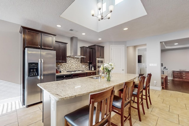 kitchen featuring appliances with stainless steel finishes, a breakfast bar, a textured ceiling, wall chimney range hood, and an island with sink