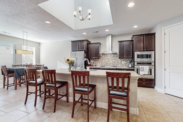 kitchen featuring a kitchen island with sink, hanging light fixtures, stainless steel appliances, and wall chimney range hood