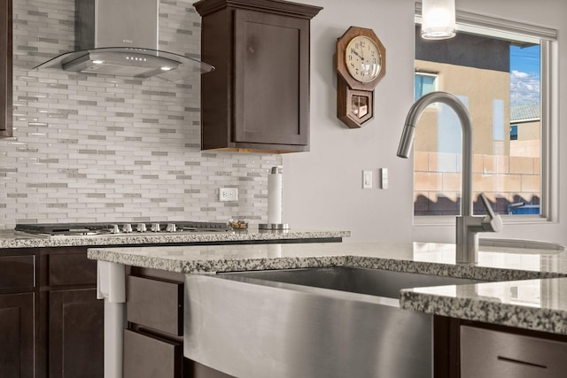 kitchen featuring wall chimney range hood, decorative backsplash, stainless steel gas stovetop, dark brown cabinets, and sink