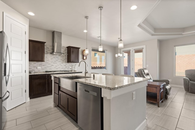 kitchen featuring a kitchen island with sink, appliances with stainless steel finishes, pendant lighting, wall chimney exhaust hood, and sink