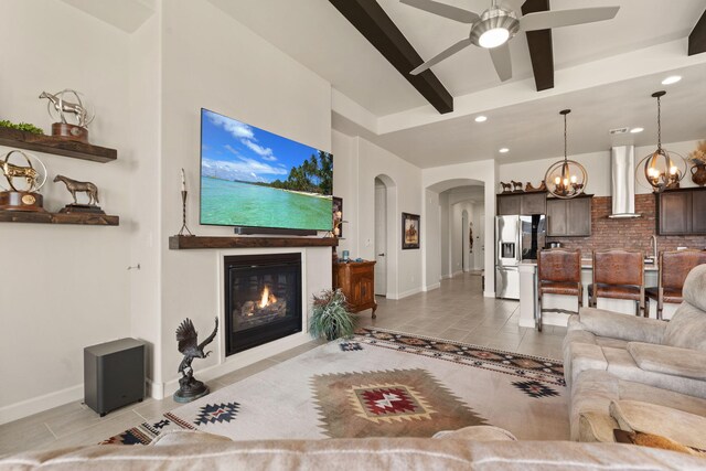 living area featuring arched walkways, beam ceiling, light tile patterned floors, a glass covered fireplace, and ceiling fan with notable chandelier