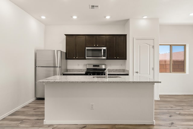 kitchen featuring sink, light wood-type flooring, light stone countertops, a center island with sink, and appliances with stainless steel finishes