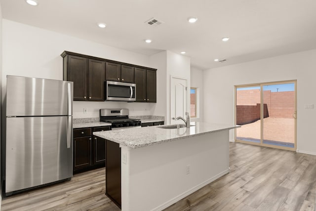 kitchen with light stone counters, stainless steel appliances, an island with sink, light wood-type flooring, and sink