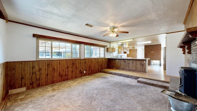 unfurnished living room featuring wood walls, light carpet, a wood stove, a textured ceiling, and ceiling fan