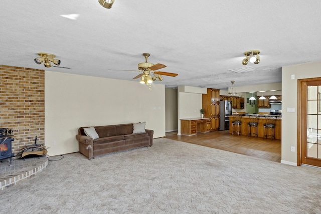 living room featuring ceiling fan, a wood stove, light colored carpet, and a textured ceiling