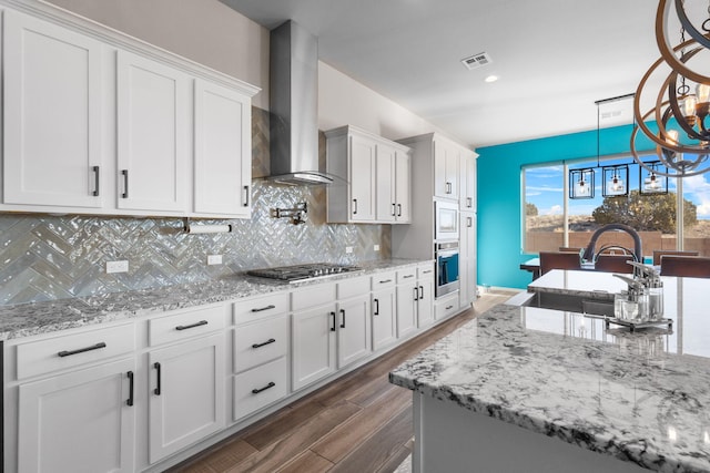 kitchen with visible vents, appliances with stainless steel finishes, hanging light fixtures, wall chimney range hood, and white cabinetry