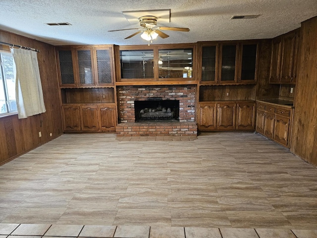 unfurnished living room with wooden walls, a brick fireplace, ceiling fan, and a textured ceiling