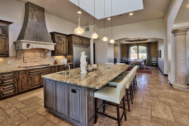 kitchen featuring decorative columns, custom range hood, stainless steel appliances, a kitchen island with sink, and a high ceiling